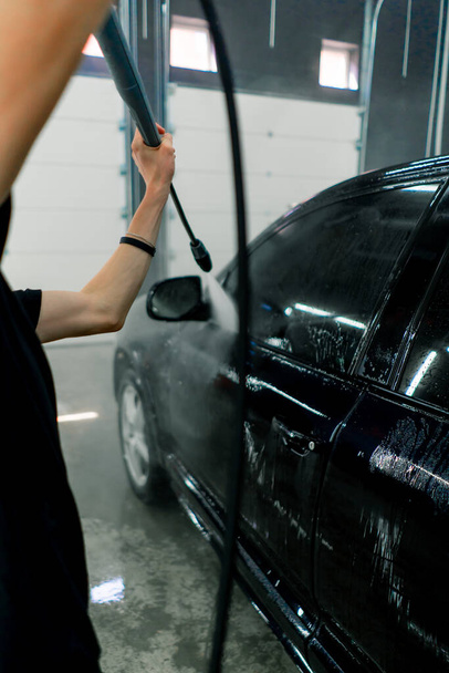 Работник автомойки моет черный роскошный автомобиль с моечной машиной высокого давления в автомойке - Фото, изображение