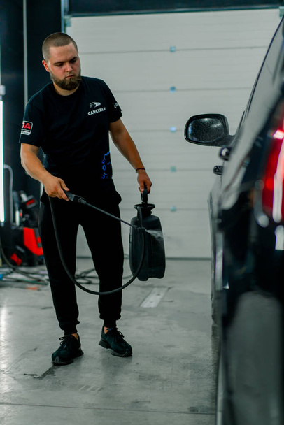 男性の洗車従業員は洗車ボックスにスプレーガンを使用して黒い高級車に洗車洗剤を適用します - 写真・画像