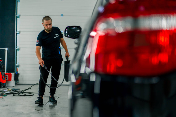 Een mannelijke carwash medewerker past carwash wasmiddel toe op een zwarte luxe auto met behulp van spuitpistool in de carwash box - Foto, afbeelding