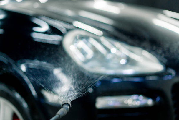 Κοντινό πλάνο του εμπρόσθιου φαναριού ενός μαύρου αυτοκινήτου κατά την εφαρμογή πλυσίματος αυτοκινήτου από μπουκάλι ψεκασμού σε κουτί πλυντηρίου αυτοκινήτων - Φωτογραφία, εικόνα