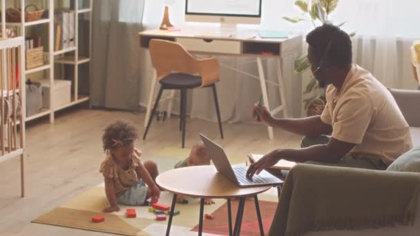 afro-americano homem no fone de ouvido com microfone trabalhando no laptop a partir de casa enquanto seus dois bonito criança crianças brincando com brinquedos nas proximidades - Filmagem, Vídeo