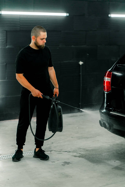 Un empleado de lavado de autos masculino aplica detergente de lavado de autos a un auto de lujo negro usando pistola en la caja de lavado de autos - Foto, Imagen