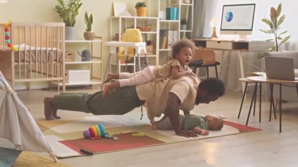 Giovane uomo afroamericano attivo e padre di due bambini piccoli che fanno flessioni a casa con la figlia sdraiata sulla schiena e il figlio tra le braccia - Filmati, video