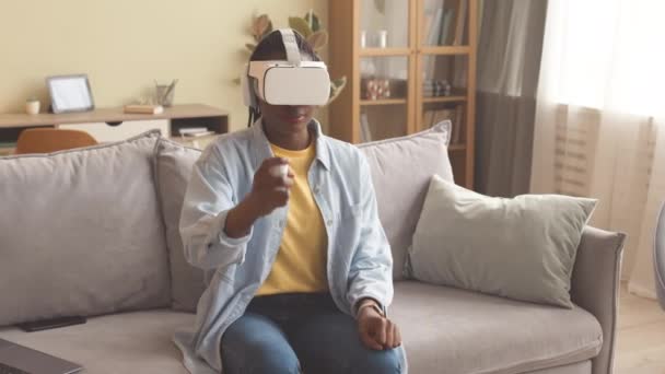 Medium shot of African American girl in vr headset sitting on couch in living room playing exciting vr game using vr gaming controller - Footage, Video