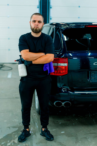 Portret van een mannelijke carwash medewerker met een spuitpistool en een microvezel doek in een garage op de achtergrond van een luxe blauwe auto - Foto, afbeelding