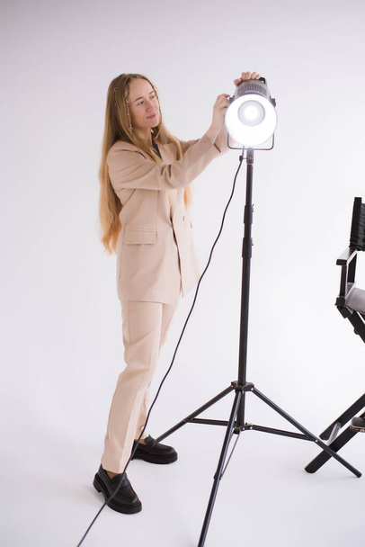 Professional female gaffer working, adjusts constant light. Woman blonde, beige suit on white isolated background in photo studio. Vertical, copy space - Фото, зображення