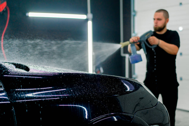 Pracownik myjni samochodowej nakłada piankę samochodową na luksusowy czarny samochód używając pistoletu natryskowego w pudełku myjni samochodowej - Zdjęcie, obraz