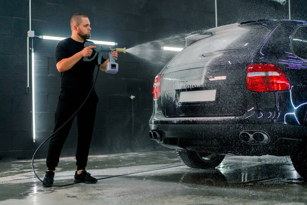 A male car wash employee applies car wash foam to a luxury black car using spray gun in the car wash box - 写真・画像