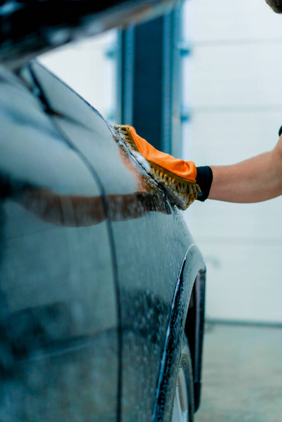 Nahaufnahme eines Autowäschearbeiters, der ein Mikrofasertuch benutzt, um ein schwarzes Luxusauto mit Autowäsche-Shampoo zu waschen - Foto, Bild