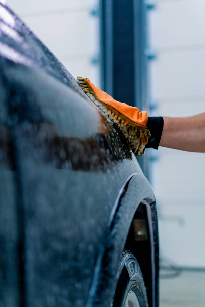 Κοντινό πλάνο ενός εργάτη πλυντηρίου αυτοκινήτων χρησιμοποιώντας ένα πανί μικροϊνών για να πλύνετε το μαύρο πολυτελές αυτοκίνητο με σαμπουάν πλυντηρίου αυτοκινήτων - Φωτογραφία, εικόνα
