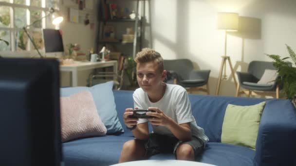 Plan long moyen d'un adolescent assis sur un canapé dans le salon et jouant à un jeu vidéo à la télévision avec excitation - Séquence, vidéo