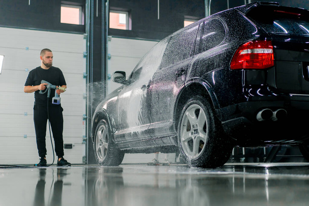 Працівник мийки чоловіків наносить піну для миття автомобілів на розкішний чорний автомобіль, використовуючи розпилювач у коробці для миття автомобілів - Фото, зображення