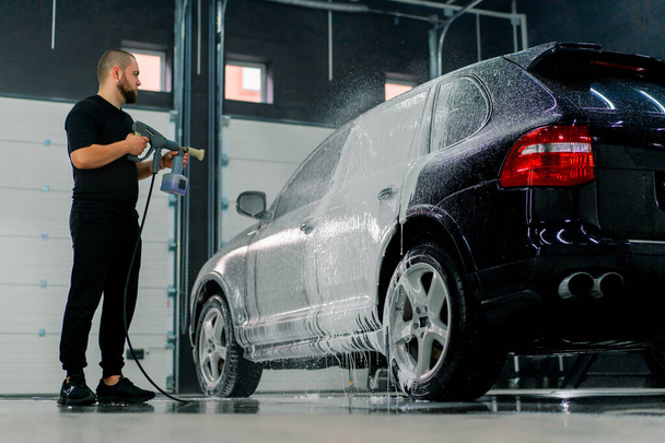 Muž mytí aut zaměstnanec aplikuje auto mytí pěny na luxusní černé auto pomocí stříkací pistole v myčce - Fotografie, Obrázek