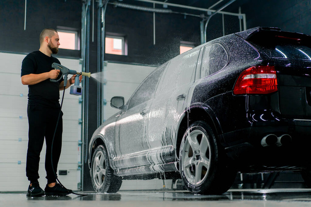 Een mannelijke carwash medewerker brengt carwash foam aan op een luxe zwarte auto met behulp van spuitpistool in de carwash box - Foto, afbeelding