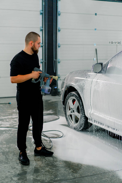 A male car wash employee applies car wash foam to a luxury black car using spray gun in the car wash box - Foto, imagen
