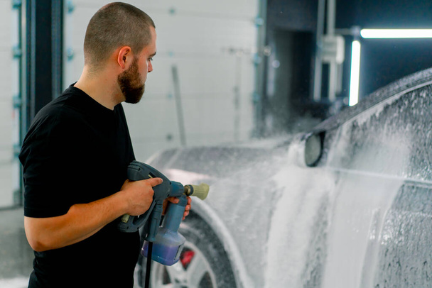 Ein Mitarbeiter einer Autowäsche sprüht mit einer Sprühpistole in der Waschbox Schaum auf ein schwarzes Luxusauto - Foto, Bild