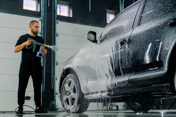A male car wash employee applies car wash foam to a luxury black car using spray gun in the car wash box - Foto, afbeelding
