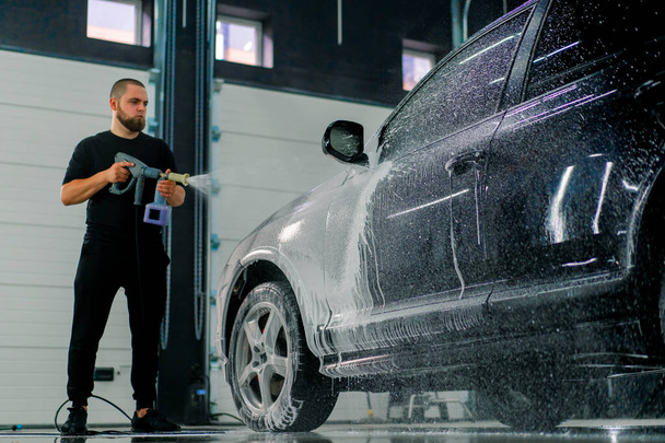 Un dipendente maschio di autolavaggio applica schiuma di autolavaggio a un'auto nera di lusso utilizzando pistola a spruzzo nella scatola di autolavaggio - Foto, immagini