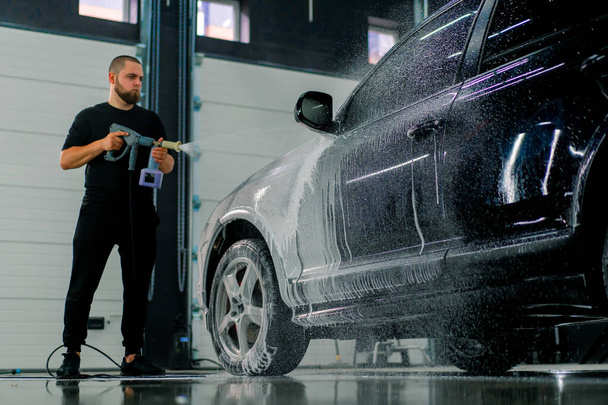 Un employé de lavage de voiture masculin applique de la mousse de lavage de voiture à une voiture noire de luxe à l'aide d'un pistolet dans la boîte de lavage de voiture - Photo, image