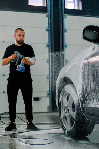 Miespuolinen autopesulatyöntekijä käyttää autopesulavaahtoa ylelliseen mustaan autoon käyttäen ruiskupistoolia autopesulaatikossa - Valokuva, kuva