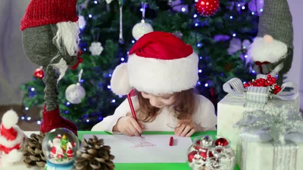 Una niña con un sombrero rojo de Santa Claus escribe una carta a Santa Claus con sus deseos bajo el árbol de Navidad. Ambiente de Navidad y Año Nuevo. Movimiento lento. - Metraje, vídeo