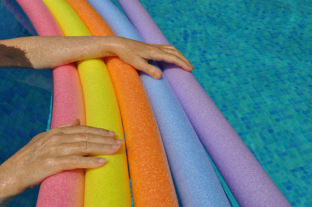 Regenboog gekleurde drijvende schuimstokken en vrouwelijke handen, in zwembad - Foto, afbeelding