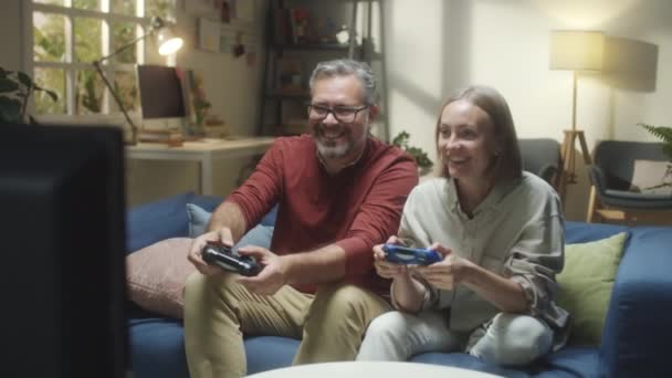 Excited wife and husband sitting on sofa in living room and playing console game together - Кадры, видео