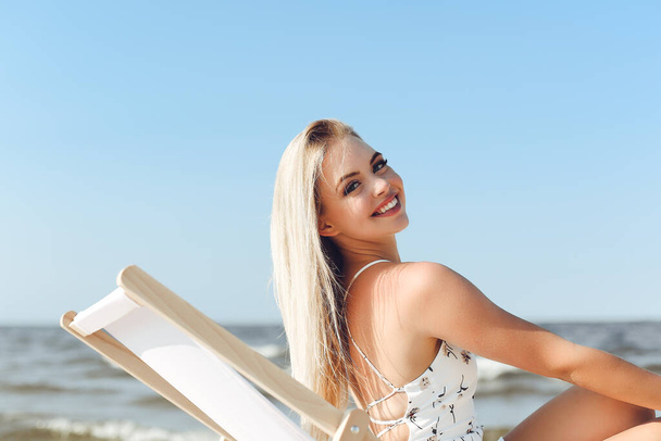 Happy blonde woman relaxing on a wooden deck chair at the ocean beach. - Zdjęcie, obraz