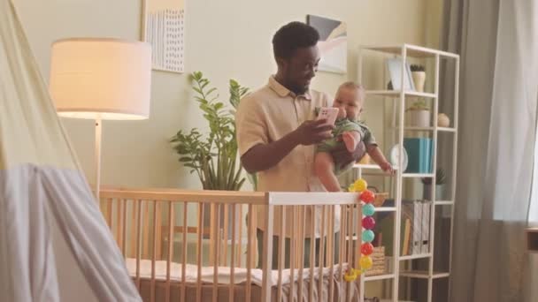 Onnellinen nuori Afrikkalainen Amerikkalainen mies käyttää älypuhelinta, kun taas rocking hänen söpö vauva poika kotona - Materiaali, video
