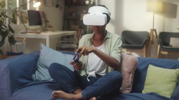Afroamerykanka w VR siedzi na kanapie i używa gamepada podczas gry w rozszerzonej rzeczywistości w domu - Materiał filmowy, wideo