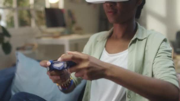 African American γυναίκα σε VR ακουστικά κάθεται στον καναπέ και χρησιμοποιώντας gamepad, ενώ παίζει το παιχνίδι στην εικονική πραγματικότητα στο σπίτι - Πλάνα, βίντεο