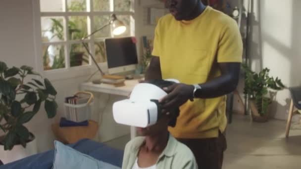 African American man helping girlfriend to put on VR headset and play game in augmented reality while spending leisure time together at home - Footage, Video