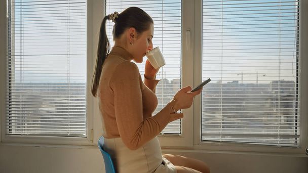 Молода бізнес-леді має кавові перерви у великому вікні за допомогою смартфона. Люди вранці, красивий міський пейзаж, відпочивають в офісі
 - Фото, зображення