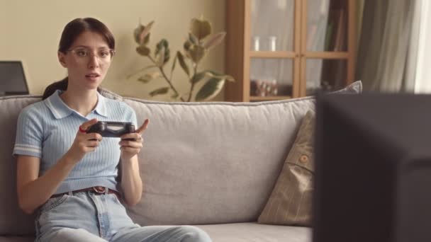 Concentrated Caucasian girl in her early 20s using controller while playing video game on sofa at home - Footage, Video