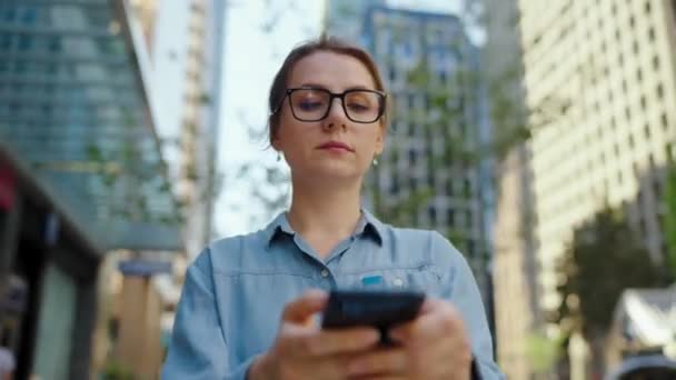 Gözlüklü beyaz kadın şehirde dolaşıyor ve akıllı telefon kullanıyor. Arka planda gökdelenler var. İletişim, iş günü, yoğun yaşam konsepti. - Video, Çekim