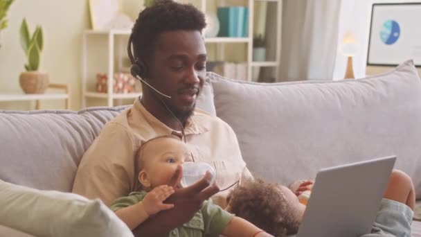 Young African American man wearing headset with microphone working on laptop from home and taking care of his two toddler kids lying on sofa nearby - Footage, Video