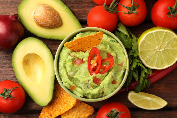 Bowl of delicious guacamole, nachos chips and ingredients on wooden table, flat lay - Zdjęcie, obraz