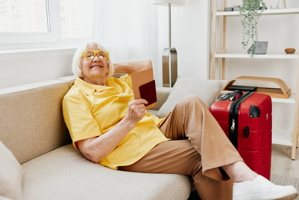 Pasaportu ve seyahat bileti olan mutlu bir son sınıf öğrencisi kırmızı bavul, tatil ve sağlık sigortası hazırladı. Gülümseyen yaşlı kadın yolculuktan önce koltukta mutlu bir şekilde oturur. Yüksek kalite fotoğraf - Fotoğraf, Görsel