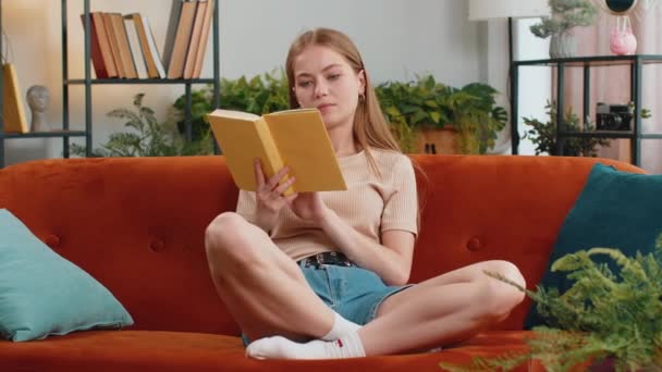 Joven mujer bonita relajante lectura interesante libro pasando páginas sonriendo disfrutando de la literatura tomando un descanso en cómodo sofá. Chica alegre pacífica en casa apartamento sala de estar sentado en el sofá - Metraje, vídeo