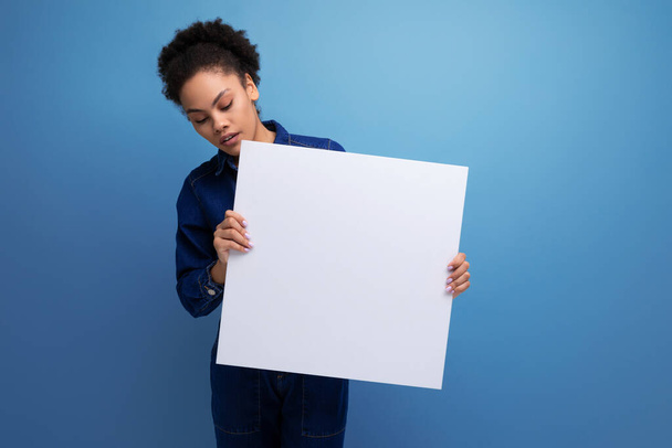 satisfied positive young hispanic brunette woman with fluffy curly hair in blue denim suit holding a white sheet of paper with a mockup for advertising. - Photo, Image