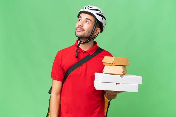 Young handsome caucasian man with thermal backpack and holding takeaway food over isolated background thinking an idea while looking up - Photo, Image