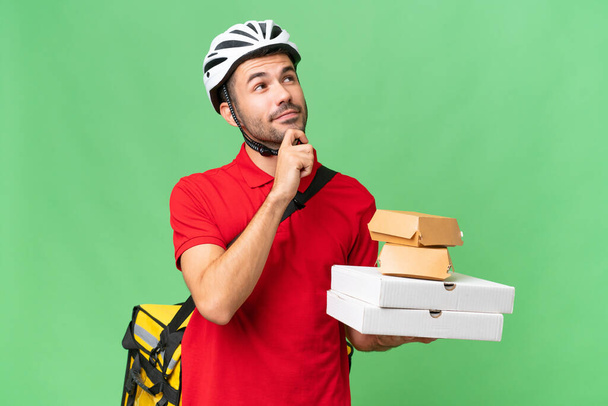 Young handsome caucasian man with thermal backpack and holding takeaway food over isolated background and looking up - Photo, Image