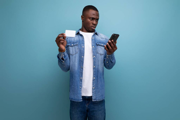 jeune américain joyeux à la peau foncée habillé élégamment en jeans utilise smartphone et carte pour faire du shopping en ligne. - Photo, image