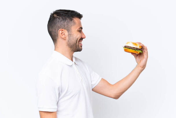 Fiatal jóképű kaukázusi férfi kezében egy burger felett elszigetelt háttér boldog kifejezés - Fotó, kép