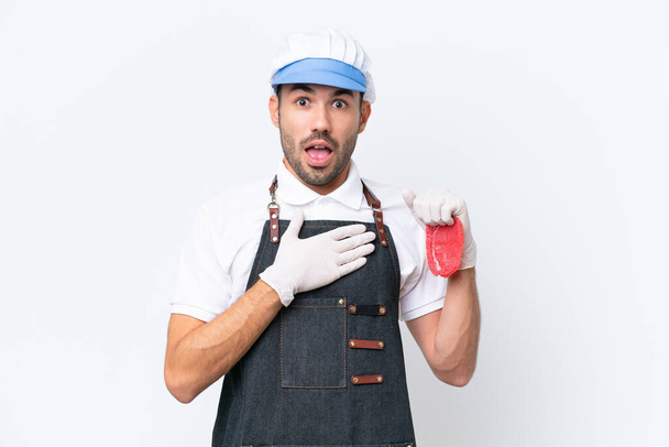Boucher homme caucasien portant un tablier et servir de la viande fraîche coupée sur fond blanc isolé surpris et choqué tout en regardant à droite - Photo, image