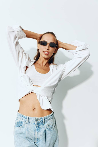 Beautiful female fashion model with tanned skin and athletic body smiling with glasses posing on white background in jeans and shirt. Body care and skin health concept with spf. High quality photo - Fotó, kép