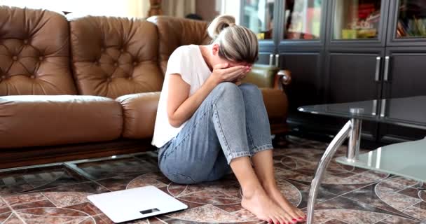 Een vrouw thuis, zittend op de vloer huilt bij de weegschaal, een close-up, slow motion. Gewichtswinst, slecht dieet - Video