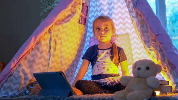 Tabletli mutlu kız çocuğu evdeki çadırda karanlıkta oturuyor. Yüksek kalite fotoğraf - Fotoğraf, Görsel