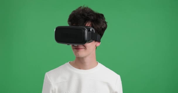 Uomo caucasico mentre indossa un visore VR su uno sfondo verde vibrante. Uomo con un sorriso contagioso e un senso di meraviglia mentre esplora un ambiente virtuale utilizzando tecnologie all'avanguardia. - Filmati, video