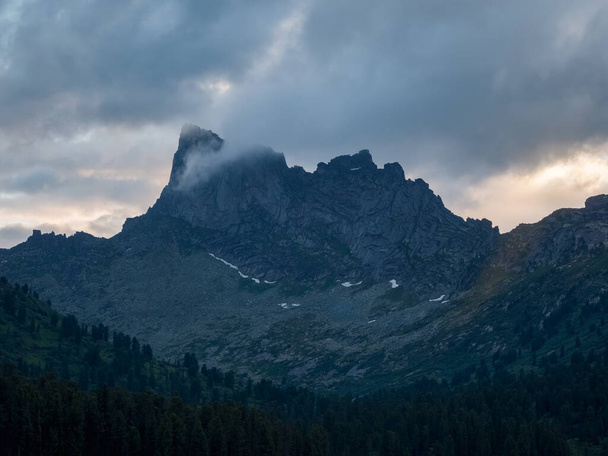Темний атмосферний сюрреалістичний пейзаж з темною скелястою вершиною гір у низьких хмарах. Сіра низька хмарність на високій плиті. Високий чорний камінь у низьких хмарах. Сюрреалістичні похмурі гори. Західних Сайянах. - Фото, зображення